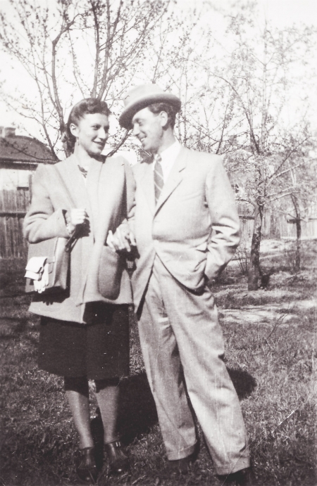 Black-and-white photograph of a man and woman, standing together, arm-in-arm, in a field outdoors with trees in the background. The couple smiles at one another and the man wears a hat and suit.
