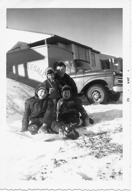 Photo en noir et blanc d'un homme avec trois jeunes enfants, assis à l'extérieur dans la neige. Ils sourient à la caméra, rassemblés ensemble devant une maison avec un camion en arrière-plan.