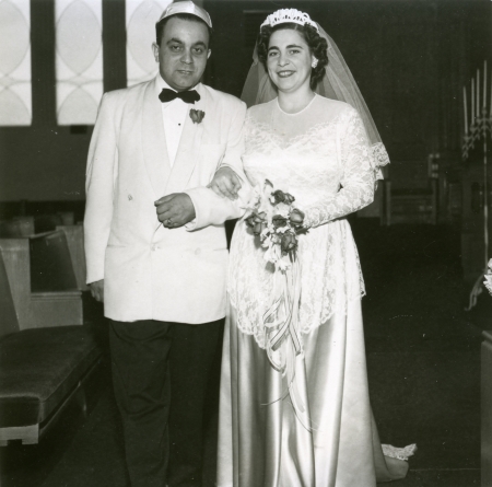 Photo en noir et blanc d'un homme et d'une femme se tenant à l'intérieur bras-dessus bras-dessous. Le couple semble célébrer leur mariage. L'homme porte un complet avec un veston de couleur claire et un nœud papillon, et la femme une longue robe blanche dont la jupe est en soie, un voile et elle tient un bouquet de fleurs.