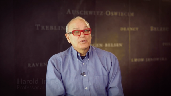 Screenshot from Harold Troper interview. He is sitting in front of a grey wall with various camps names written on it. His face and shoulders are visible at the camera.