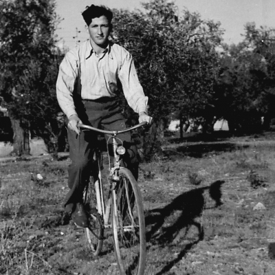 Black-and-white photograph of a young man riding a bicycle in a field. He wears dark trousers and a light collared shirt. There are trees in the background.