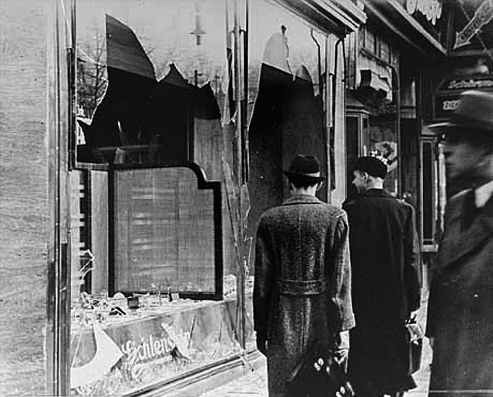 Black-and-white photograph of three men in hats and jackets, standing on a sidewalk and looking at two shattered storefront windows. Two men have their back to the camera.