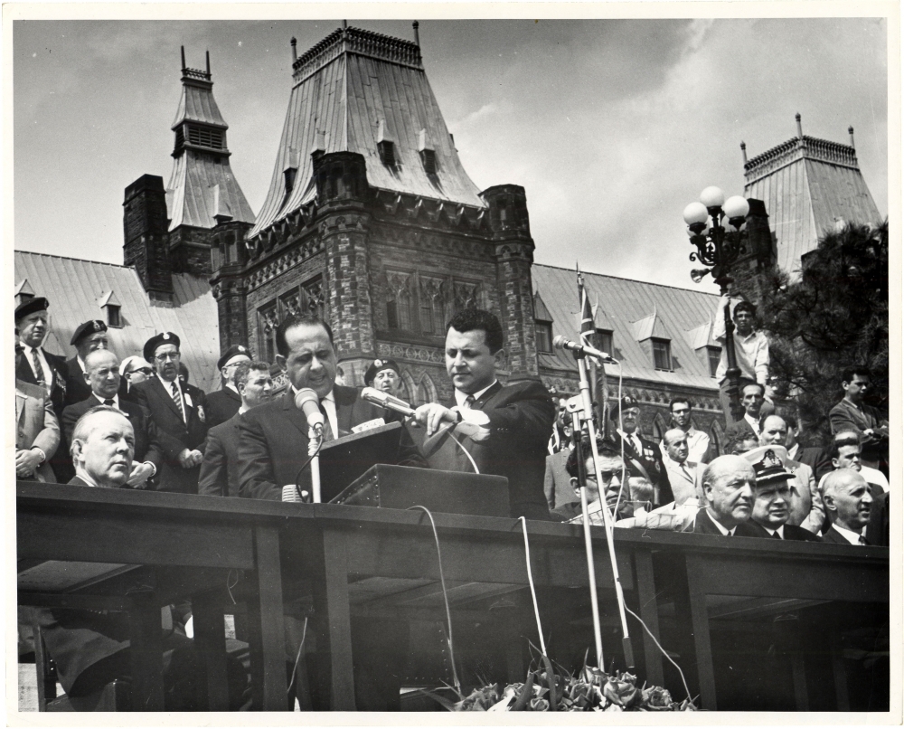 Black-and-white photograph of a man in a suit speaking into two microphones at a podium outdoors. He is surrounded by a group of people, some sitting, with a large building in the background.