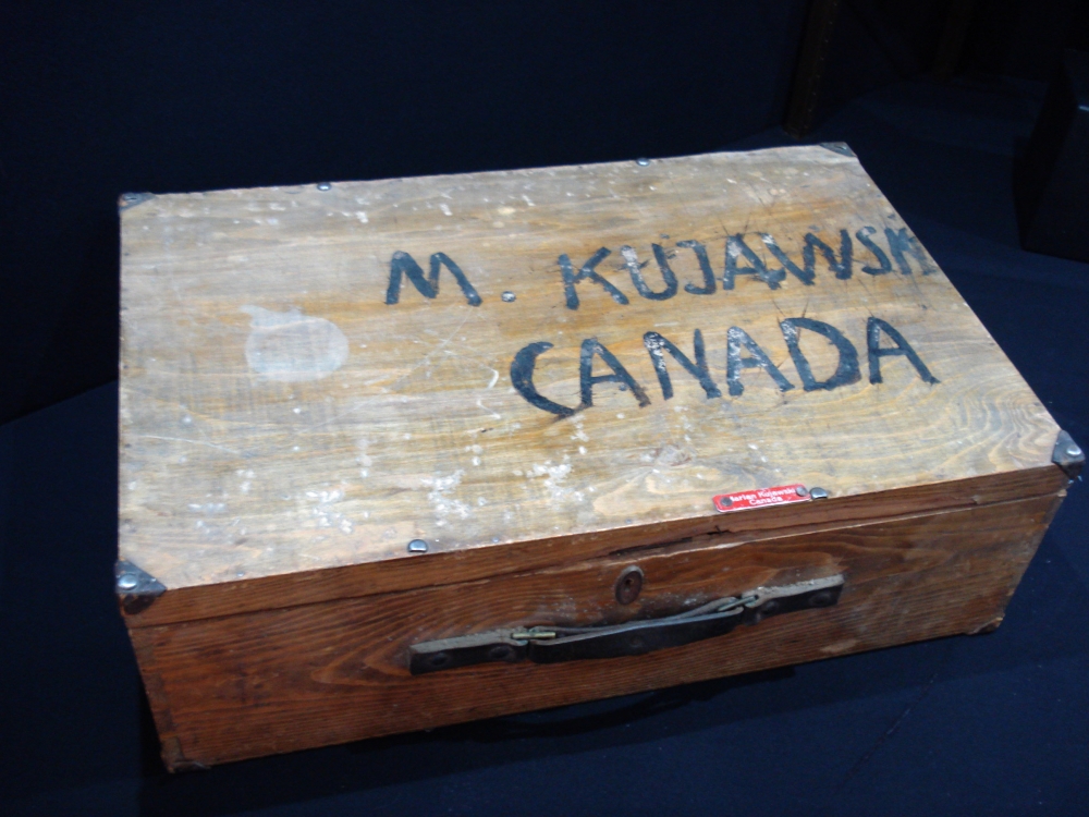 Photo en couleur d'une valise en bois de forme rectangulaire avec les mots “M KUJAWSK/CANADA” peints sur le dessus de la valise.