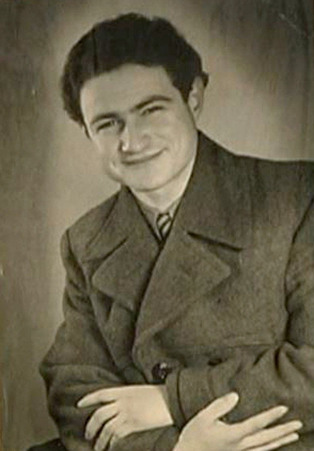 Portrait photographique d'un jeune homme, pris à partir de la taille, assis les bras croisés. Il porte un manteau et sourit à la caméra.