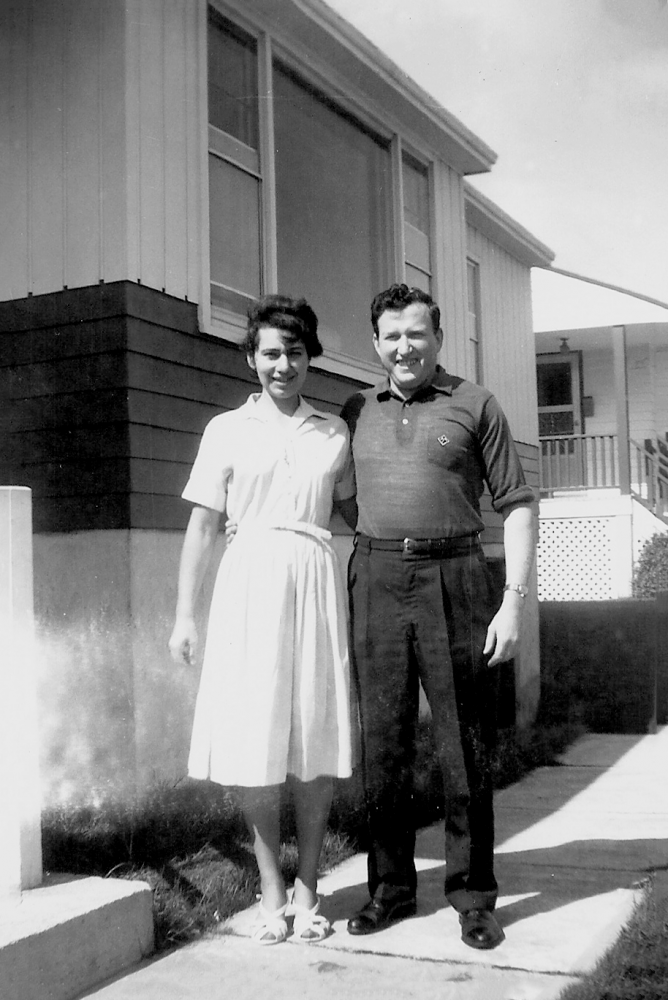 Black-and-white photograph of a man and woman smiling and standing outside the entrance to a house. It appears to be summer. The woman wears a short-sleeved dress.