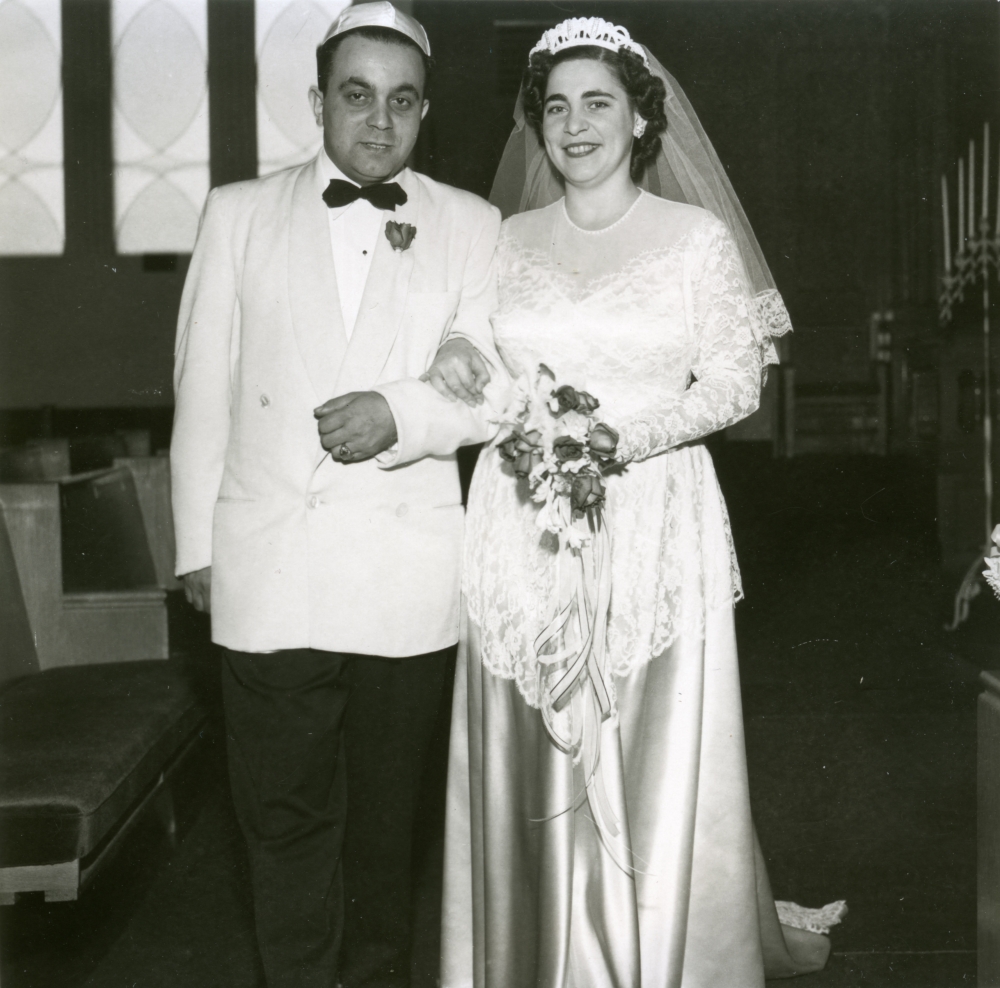 Photo en noir et blanc d'un homme et d'une femme se tenant à l'intérieur bras-dessus bras-dessous. Le couple semble célébrer leur mariage. L'homme porte un complet avec un veston de couleur claire et un nœud papillon, et la femme une longue robe blanche dont la jupe est en soie, un voile et elle tient un bouquet de fleurs.