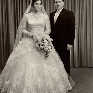 Black-and-white studio portrait photograph of a man and a woman on their wedding day. The woman wears a long white gown with a veil, holding a bouquet of flowers, and the man wears a formal suit and a moustache.