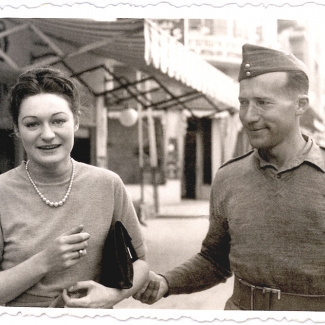 Photo en noir et blanc d'une femme et d'un homme à l'extérieur. La femme tient une petite bourse sous son bras et fait face à la caméra en souriant. L'homme regarde la femme et lui tient le coude.