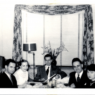 Photo en noir et blanc d'un groupe d'environ cinq adultes assis à une table à manger sur laquelle il y a des verres de vins, souriant à la caméra. Les hommes portent des complets.