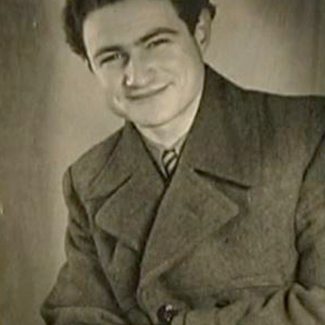 Black-and-white portrait photograph of a young man, pictured from the waist up, sitting with his arms crossed. He wears a jacket and smiles at the camera.