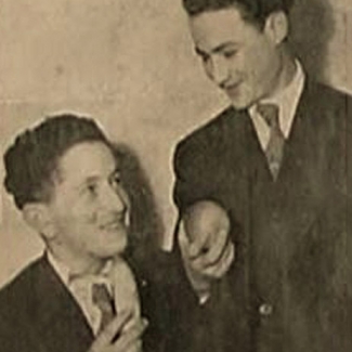 Photo en noir et blanc de deux jeunes hommes, l'un des deux est assis, qui se sourient l'un à l'autre. Les deux jeunes hommes portent des complets et tiennent ce qui semble être des morceaux de fruits.