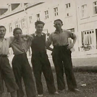 Photo en noir et blanc d'un groupe de quatre garçons se tenant bras-dessus bras-dessous sur la partie ombragée d'une route. Il y a un grand édifice de l'autre côté de la rue.
