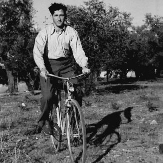 Black-and-white photograph of a young man riding a bicycle in a field. He wears dark trousers and a light collared shirt. There are trees in the background.