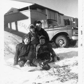 Black-and-white photograph of man with his three young children, sitting in the snow outdoors. They smile at the camera, grouped together in front of a house with a truck in the background.