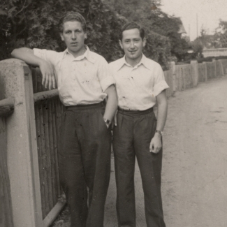 Photo en noir et blanc de deux jeunes hommes se tenant ensemble sur une route, l'un d'eux s'appuie sur une clôture. Ils portent des chemises blanches à manches courtes avec un col et des pantalons de couleur foncée.